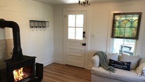 A cozy living room with a wood-burning stove, a white door, a window with stained glass, and a striped sofa.