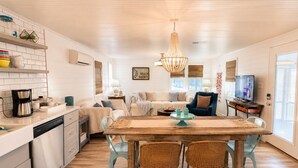 Une cuisine moderne avec des meubles blancs, un coin repas avec une table en bois et des chaises en osier, un salon avec un canapé et un téléviseur, et une grande fenêtre qui laisse entrer la lumière naturelle.