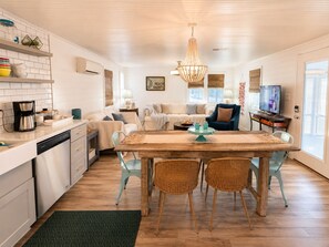 A modern kitchen with white cabinetry, a dining area with a wooden table and wicker chairs, a living room with a sofa and a television, and a large window providing natural light.