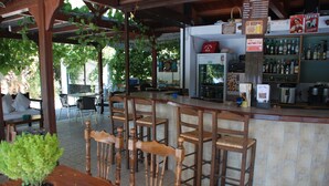 Una zona de bar con sillas de madera y un mostrador, rodeada por un porche cubierto con plantas colgantes.