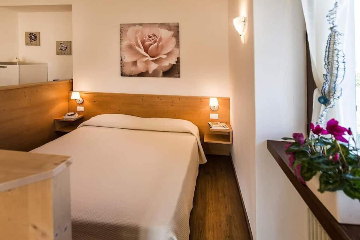 A hotel room with a wooden headboard, a single bed, bedside tables with lamps, a large floral artwork on the wall, and a potted plant by the window.
