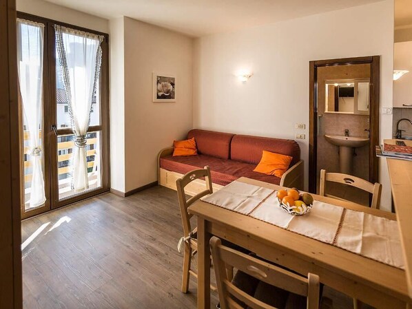 A cozy living area with a sofa, a dining table with chairs, a small kitchen area, and a sliding glass door leading to a balcony. - Le Servite Apartments (Arco)
