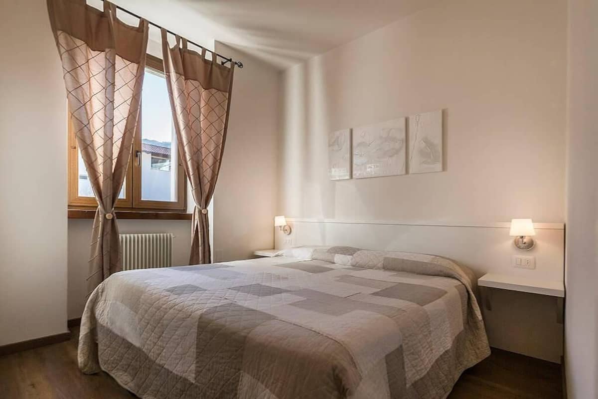 A bedroom with a large bed, a window with curtains, a bedside table with a lamp, and artwork on the wall.