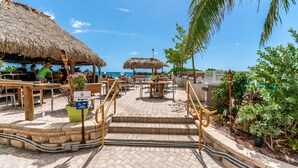 A beachside restaurant with outdoor seating, a pool access sign, and a thatched-roof structure.