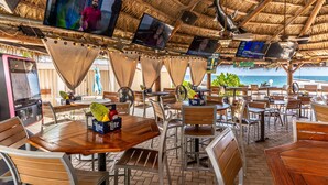 An outdoor dining area with multiple tables and chairs, screens displaying content, and a view of the ocean.