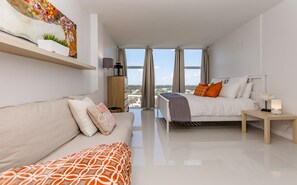 Ein modernes Hotelzimmer mit einem großen Bett, einer Couch, einem kleinen Tisch und Blick auf die Stadt durch große Fenster.