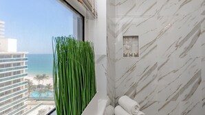 A bathroom with a view of the beach, a white marble wall, and a window with green plants.