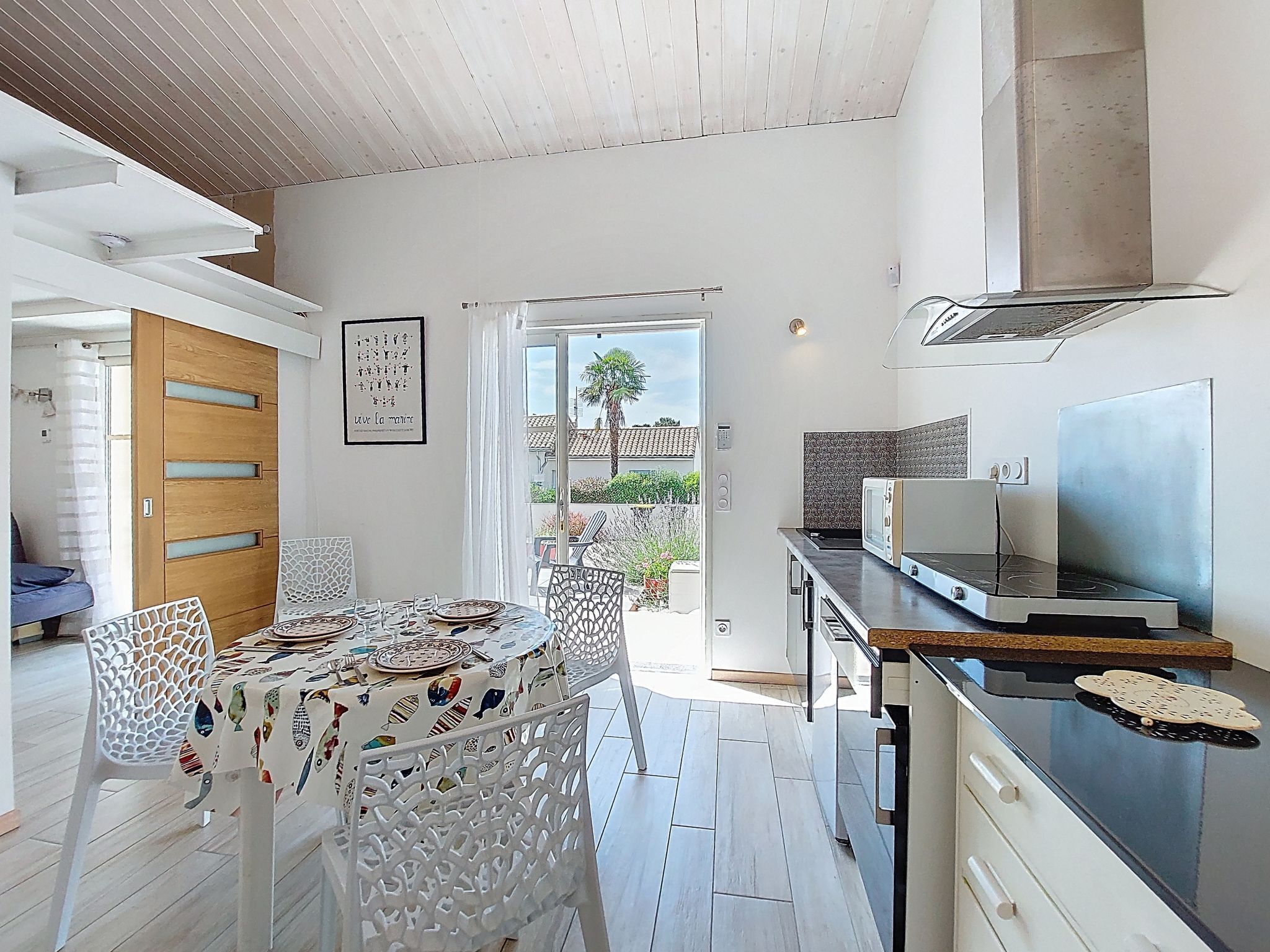 A modern kitchen with a dining area, featuring a wooden door, a framed artwork, and a view of the outdoors.