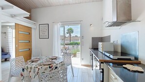 A modern kitchen with a dining area, featuring a wooden door, a framed artwork, and a view of the outdoors.