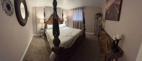 A bedroom with a four-poster bed, a nightstand with a lamp, a dresser with candles, and a painting on the wall.