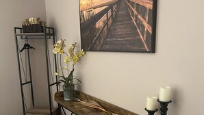 A hallway with a wooden bench, a painting of a sunset over a bridge, a plant with yellow flowers, and a candle holder with candles.