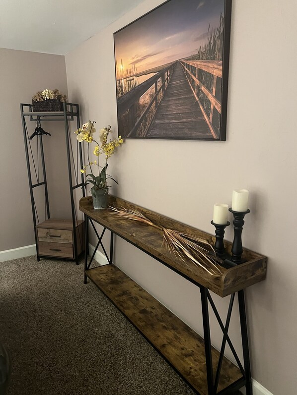 A hallway with a wooden bench, a painting of a sunset over a bridge, a plant with yellow flowers, and a candle holder with candles.