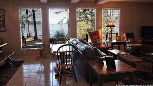Un salon comprenant un canapé, des fauteuils, une table à manger et un téléviseur. Il y a trois fenêtres qui laissent entrer la lumière naturelle.
