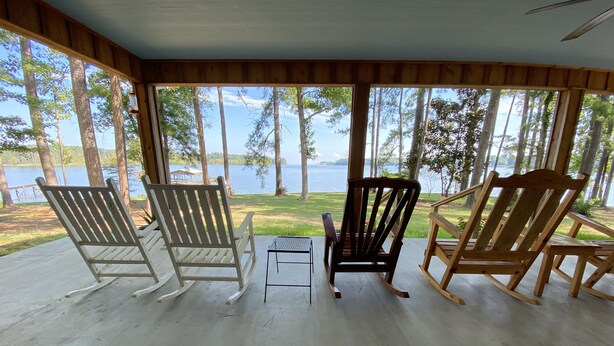 Une véranda avec trois fauteuils à bascule et une petite table, donnant sur un lac et une forêt.