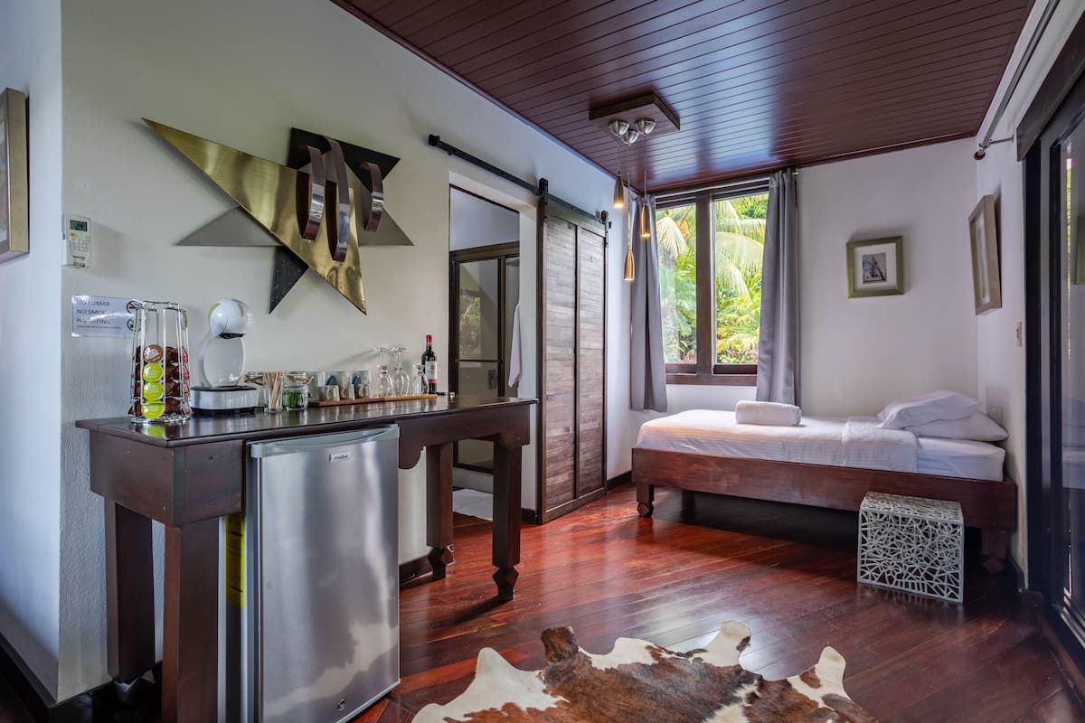 A compact room with a bed, a small kitchenette, and a large window offering a view of greenery.