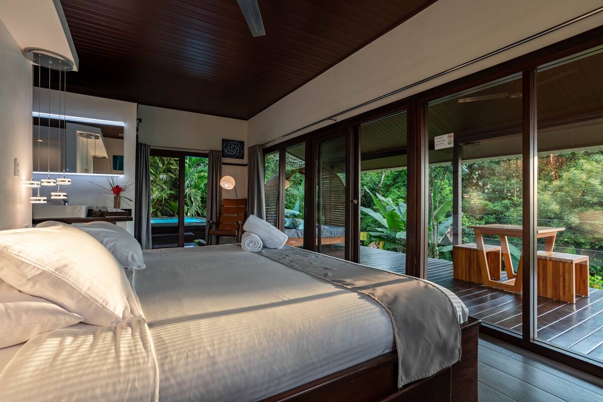 A modern bedroom with a large bed, a desk, and a view of lush greenery through large sliding doors.