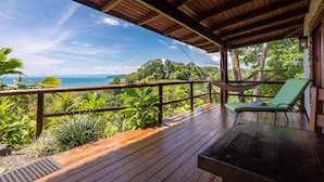 Um deck de madeira com uma rede e uma cadeira acolchoada verde, com vista para uma paisagem exuberante e o oceano.