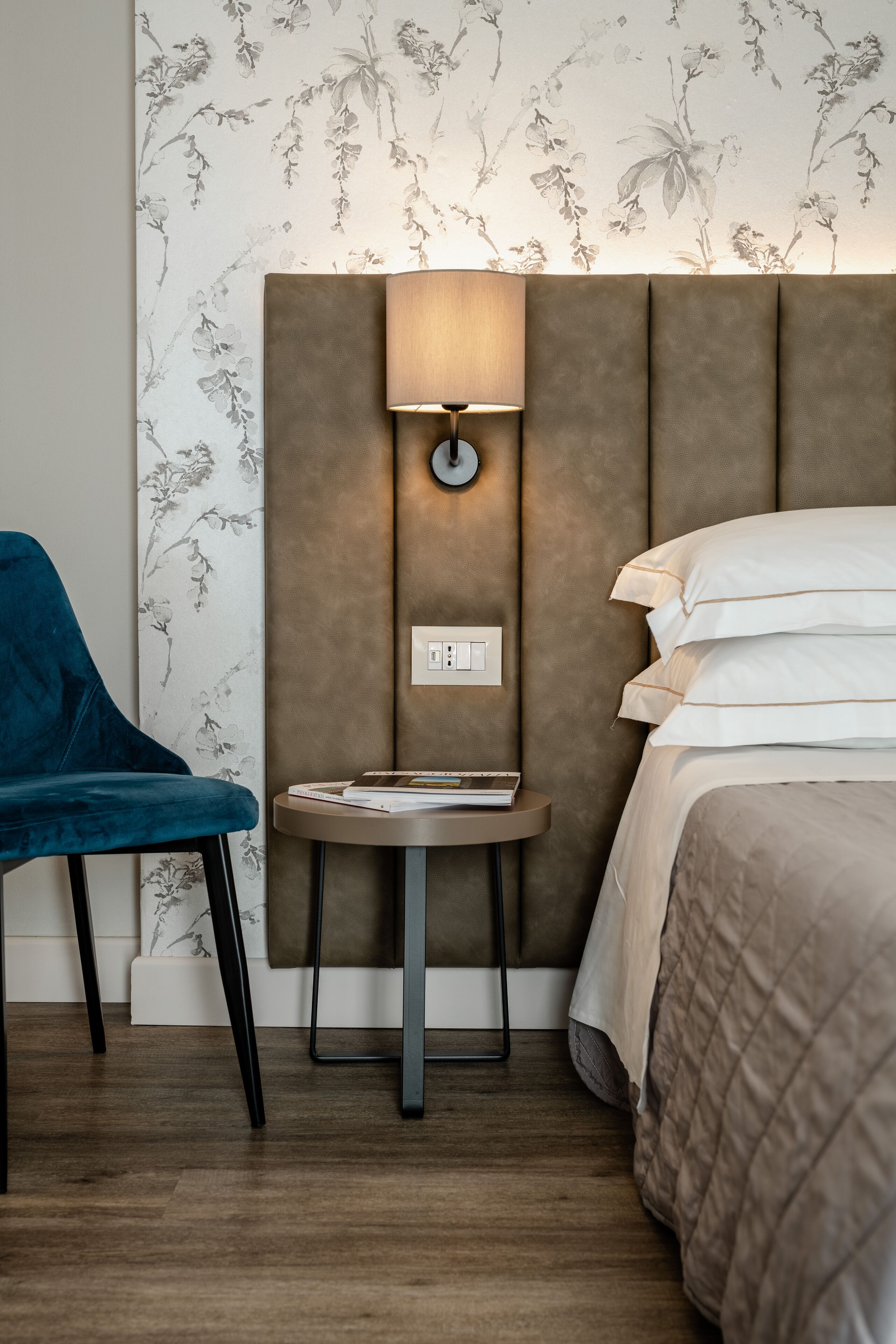Une chambre d’hôtel avec un lit, une chaise bleue, une petite table avec une lampe et un papier peint à motifs floraux.