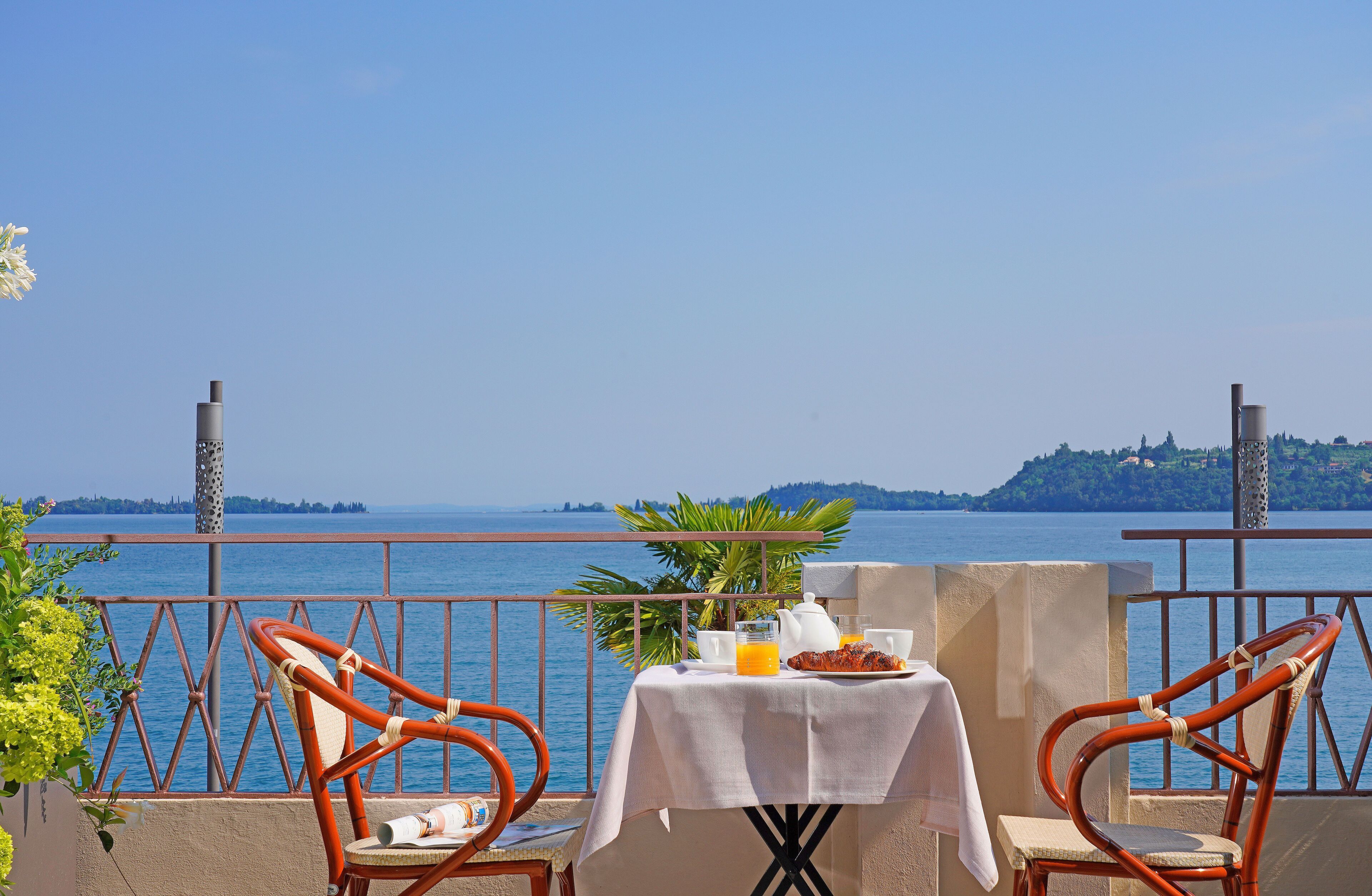 A terrace with two wicker chairs, a table with breakfast items, and a view of the sea and distant hills.