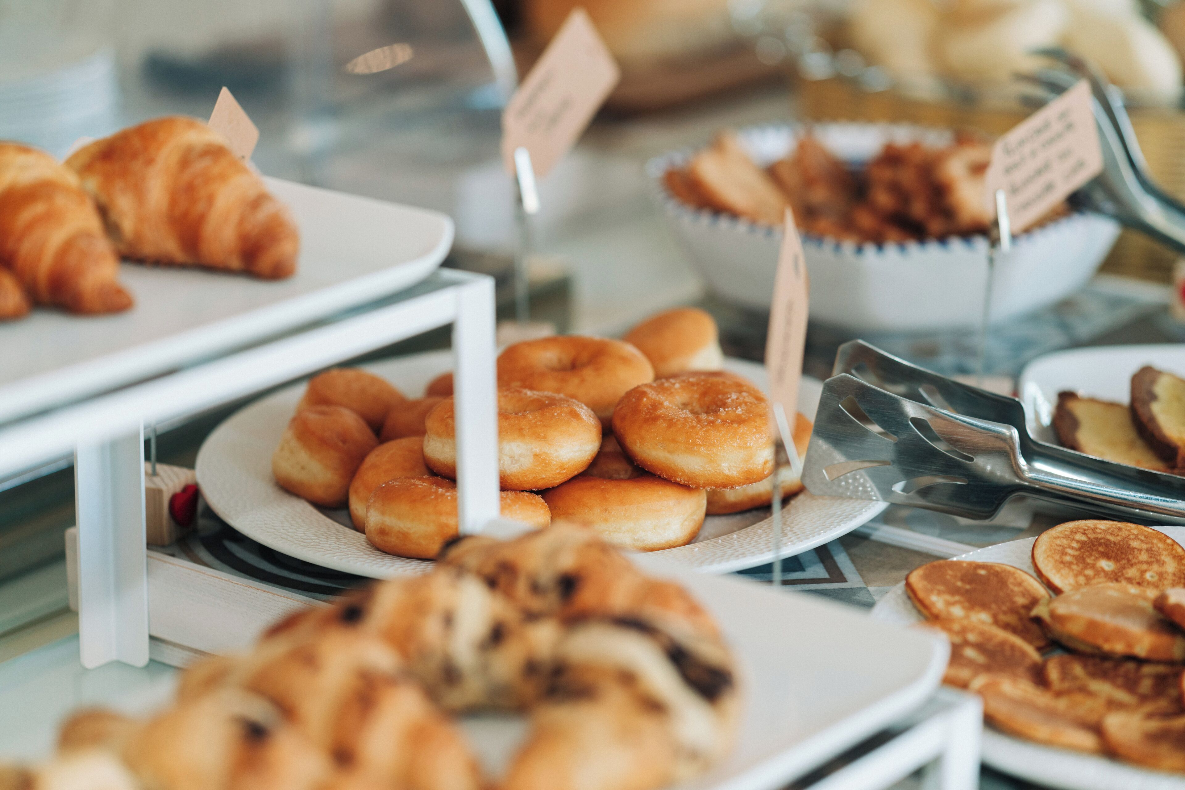 Variedad de productos horneados expuestos en platos y bandejas, como cruasanes, donas y otras exquisiteces.