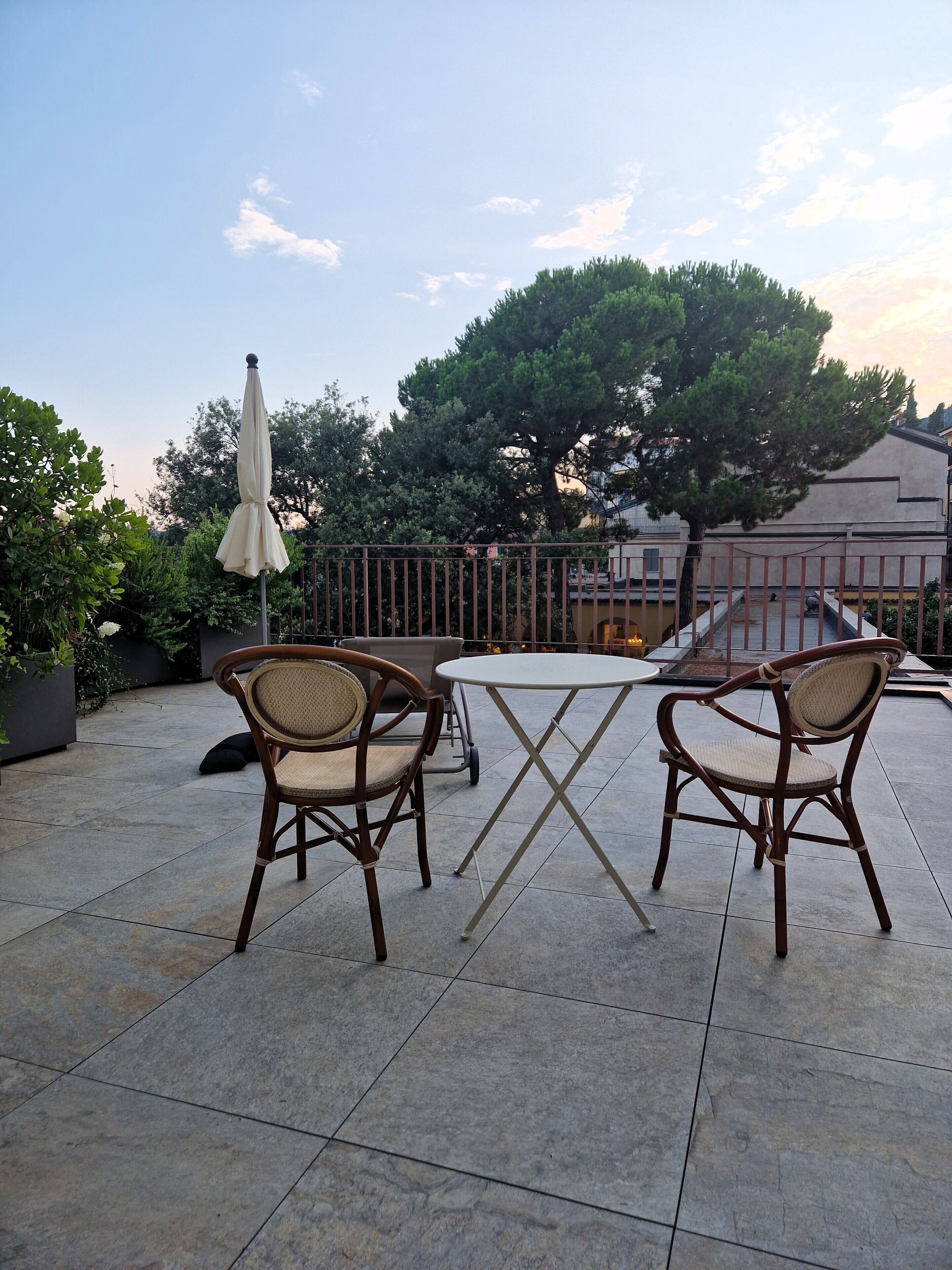 Une terrasse extérieure avec deux fauteuils en osier et une table, entourée d’une clôture en métal, avec des arbres et des bâtiments en arrière-plan.