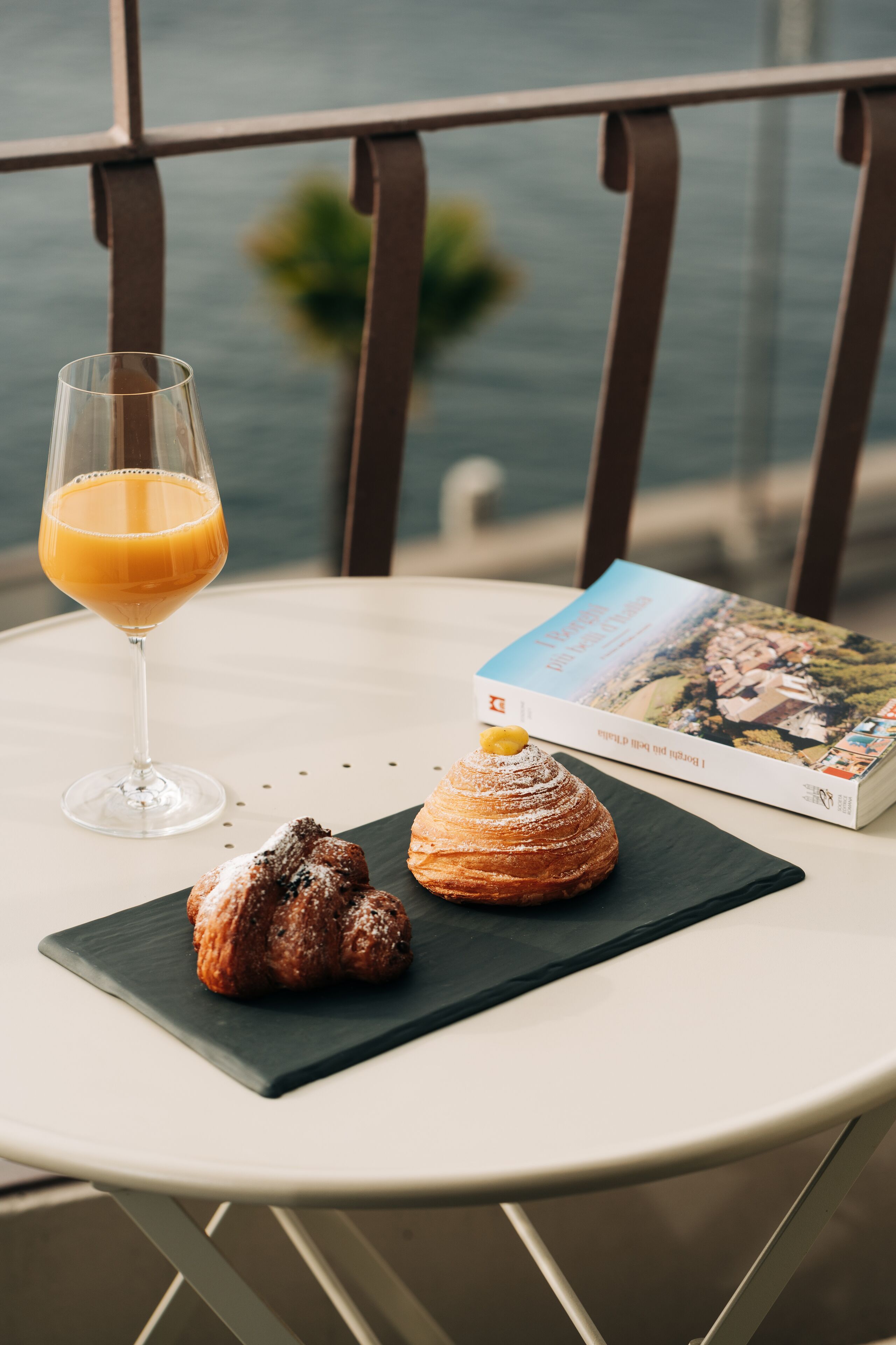 A table with a glass of orange juice, a pastry, and a book on a balcony overlooking a body of water.