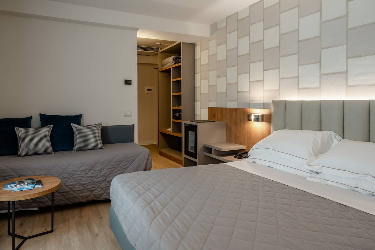 A hotel room with two beds, a small table, a bedside table, a wardrobe, and a wall-mounted light.