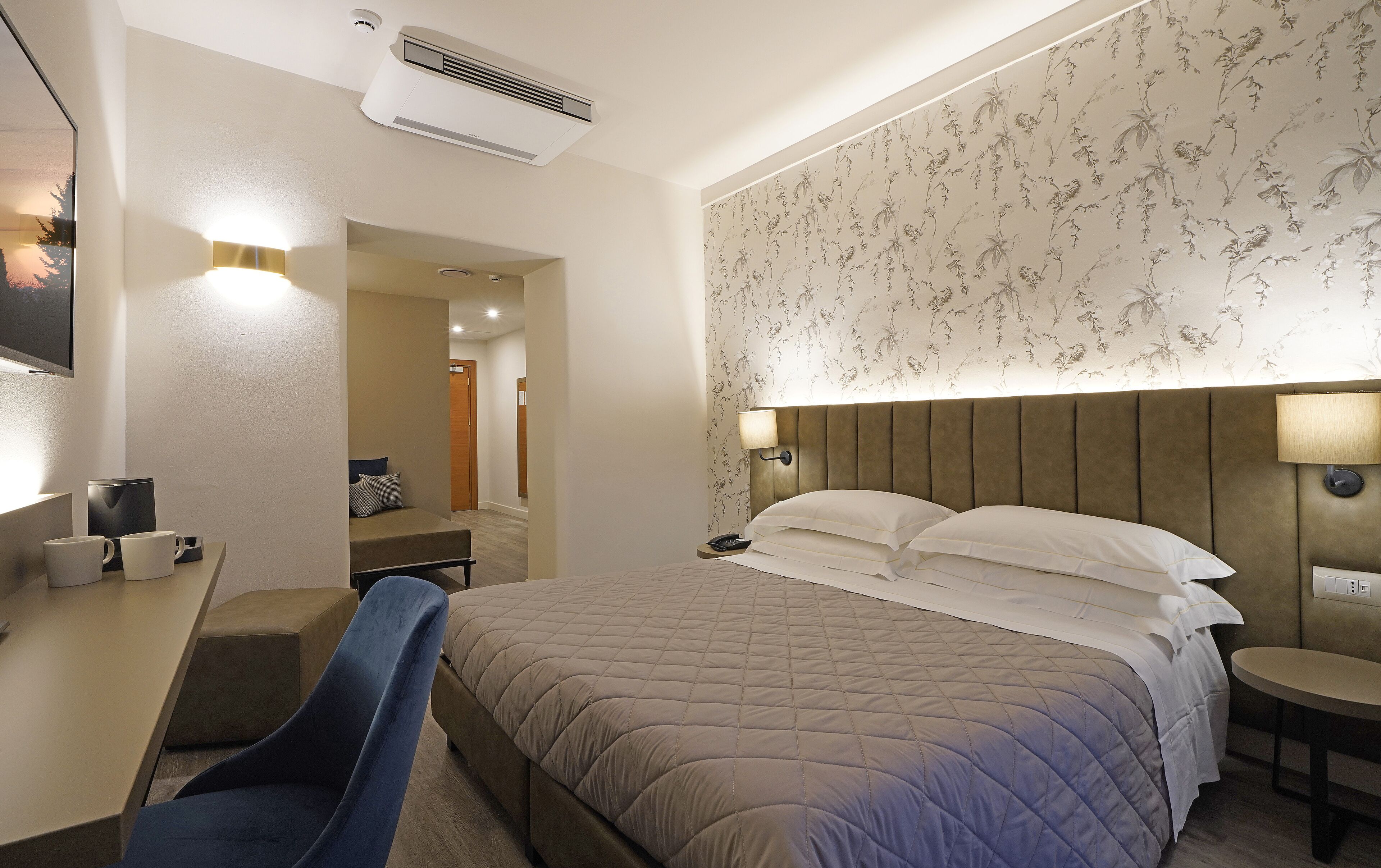 A hotel room with a bed, a desk with a chair, a TV, and a wall-mounted lamp.
