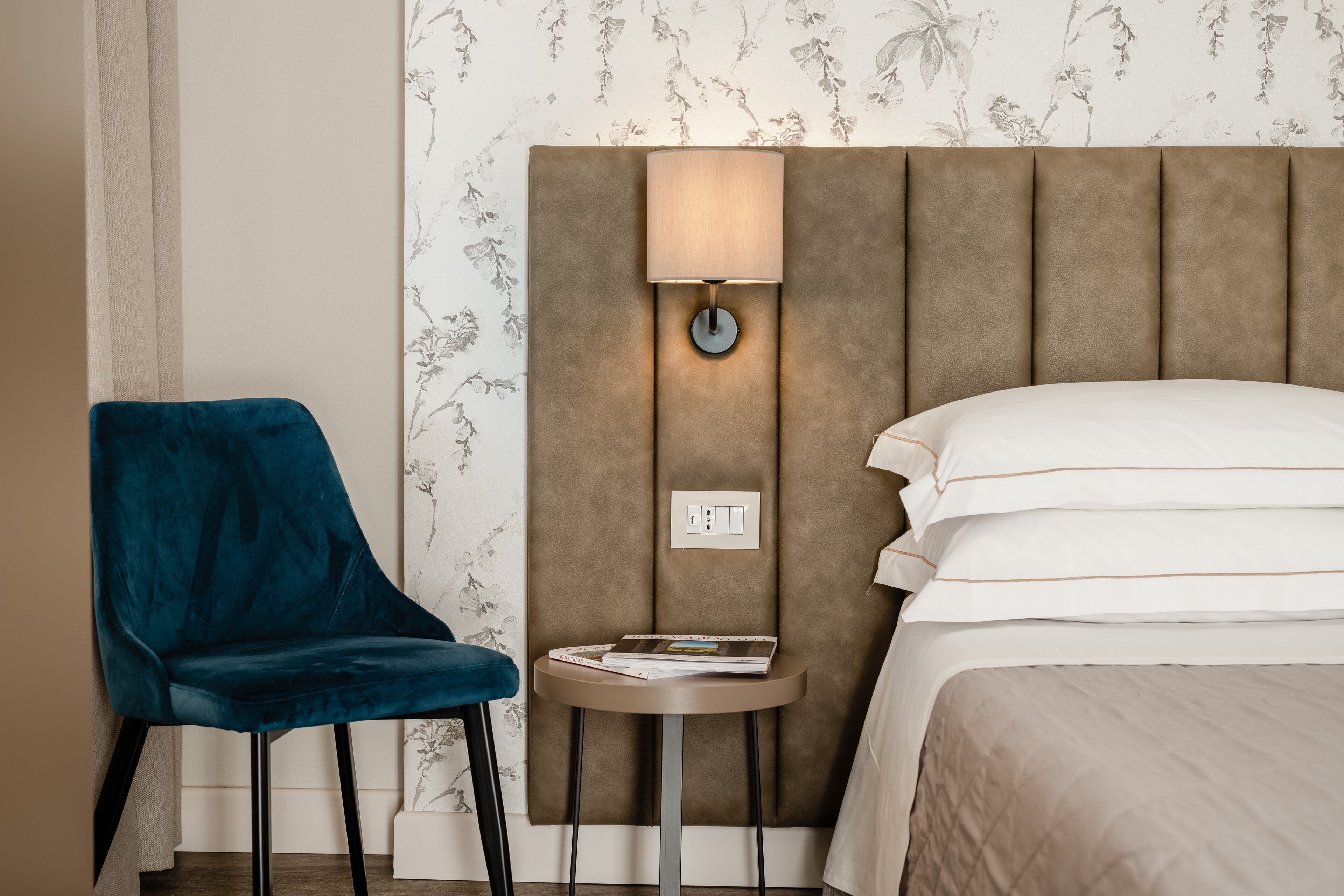 Una habitación de hotel con una cama, una silla azul, una mesita con una lámpara y una pared con papel tapiz floral.