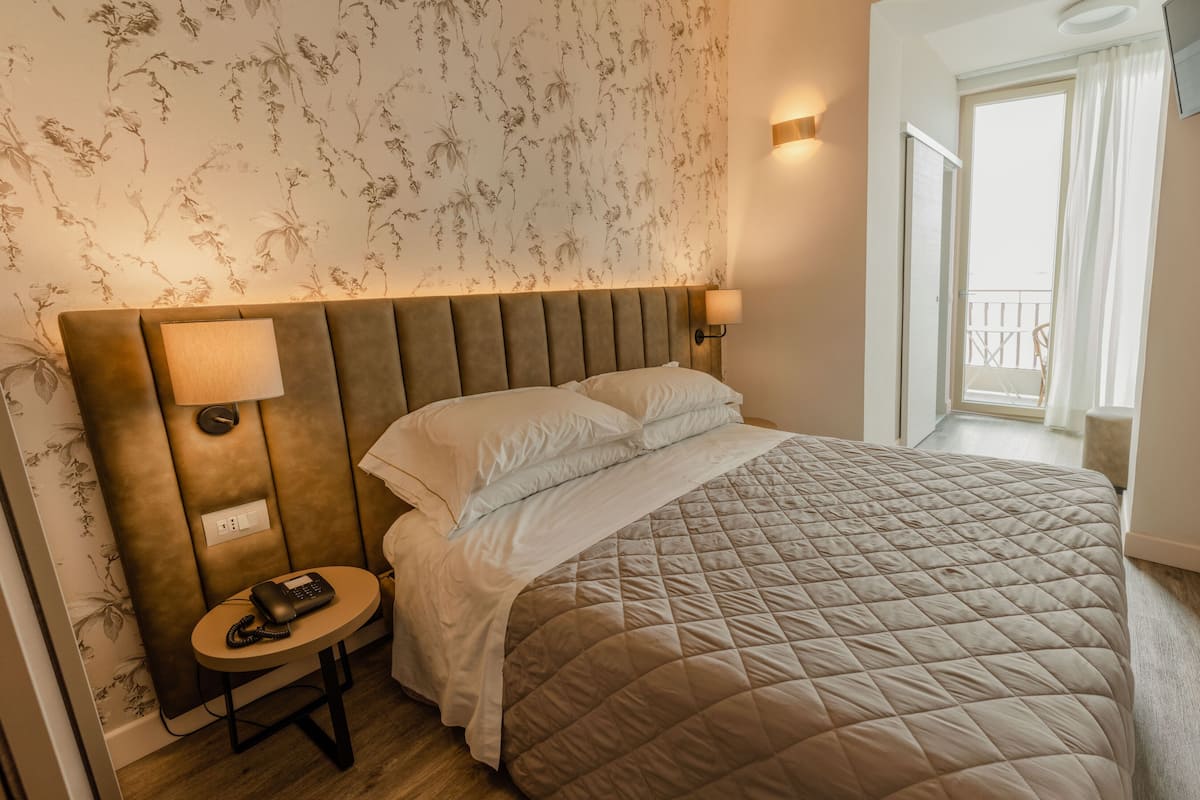 A hotel room with a large bed, a bedside table with a phone, a wall-mounted TV, and a door leading to a balcony.