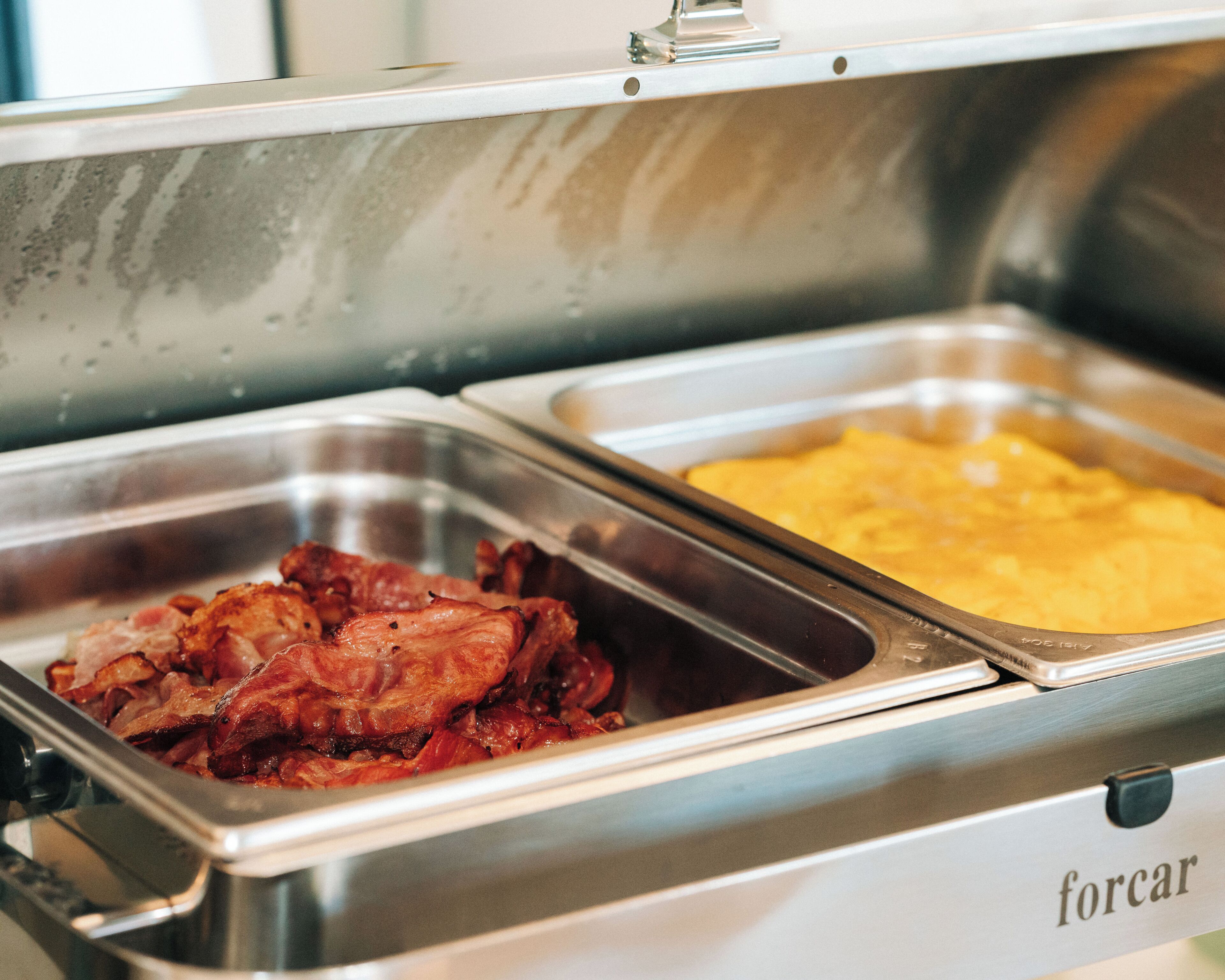 A buffet setup with two metal serving trays, one containing bacon and the other containing scrambled eggs.