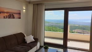 A living room with a brown sofa, a painting of a sunset, and a large window with a view of greenery and hills. - "Nilaria" sea view apartment & pool near Krk, Rijeka (Bakar)