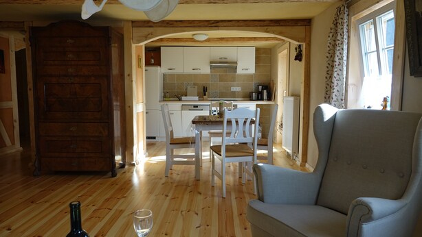 A cozy living room with a wooden floor, a grey armchair, and a dining table with chairs. A kitchen is visible in the background with white cabinets and a stove.