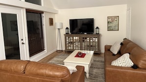 A living room with a brown leather sofa, a white coffee table, a TV on a wooden cabinet, and a large window with blinds.