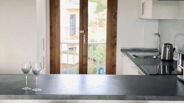 A kitchen with a dark countertop, two white chairs, and a view of a balcony through a wooden door. - Licensed Beautiful large 2 Bed Apartment in Puerto Pollenca. 2 pools and balcony (Puerto de Pollenca)