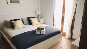 A neatly arranged bedroom with a bed, pillows, a bedside table, and a window with curtains. - Licensed Beautiful large 2 Bed Apartment in Puerto Pollenca. 2 pools and balcony (Puerto de Pollenca)