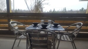 A table set with bowls and glasses on a metal table, surrounded by wicker chairs, with a wooden bench in the background and a view of trees outside.
