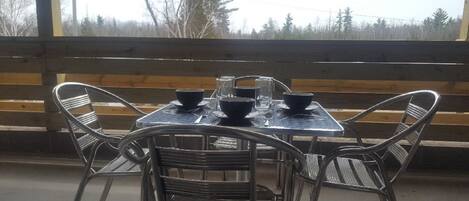 A table set with bowls and glasses on a metal table, surrounded by wicker chairs, with a wooden bench in the background and a view of trees outside.