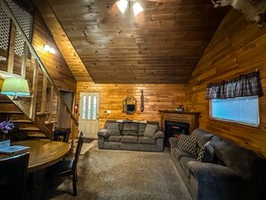 A cozy cabin interior with a wooden ceiling, a fireplace, a couch, and a dining area with a table and chairs.