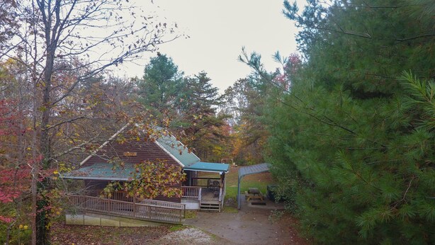 A rustic cabin with a porch and a covered picnic area, surrounded by trees and a dirt path.