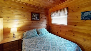 Ein Zimmer einer Holzhütte mit Bett, Nachttisch, Lampe, einem Fenster mit Jalousien und einem Wandbild.