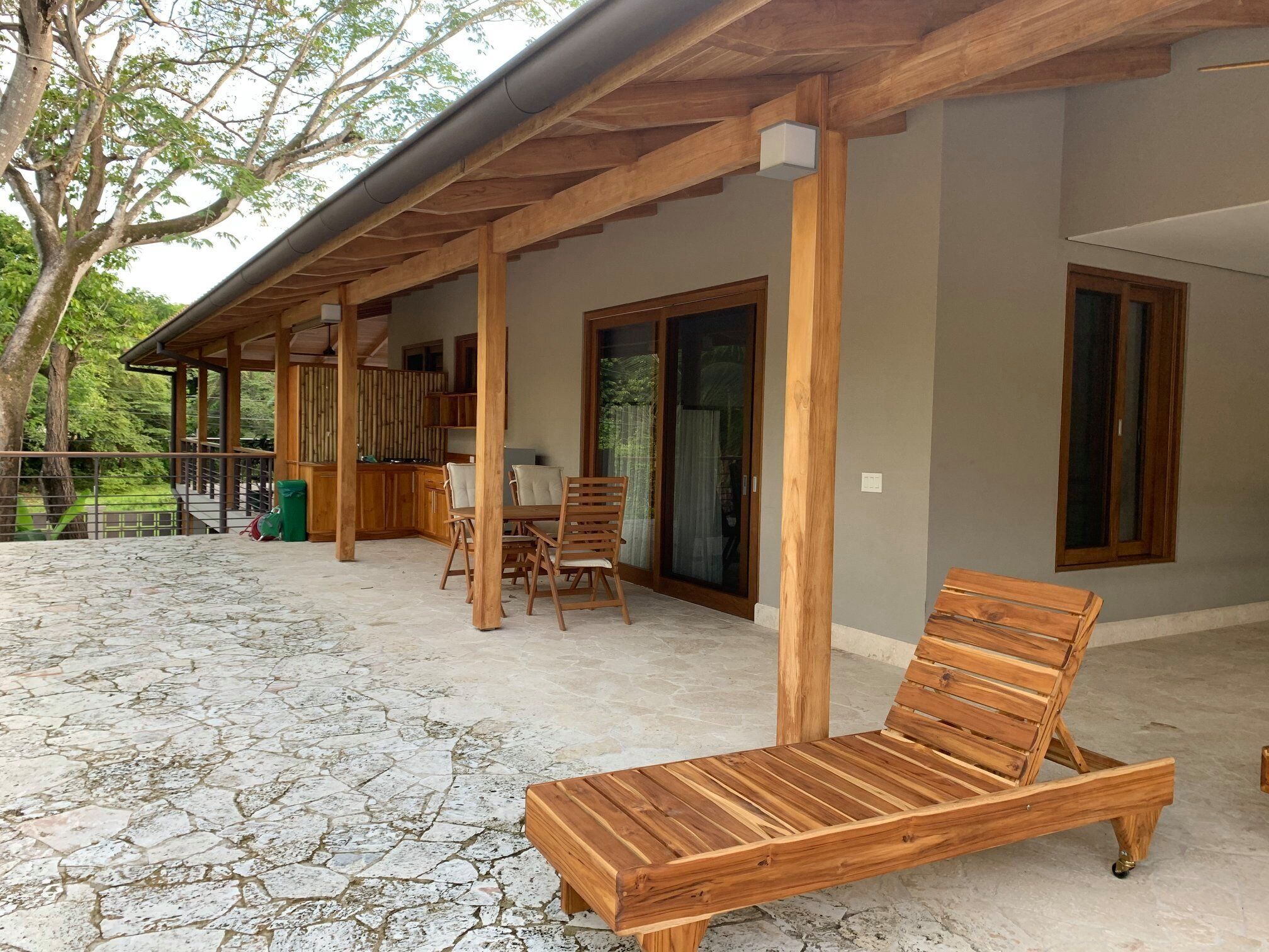 A covered outdoor area with a wooden lounge chair, a table, and chairs.