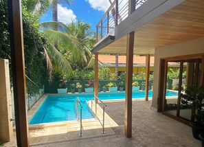 Un área de piscina con una piscina azul cristalina, rodeada de plantas en macetas y palmeras, bajo una pérgola de madera.