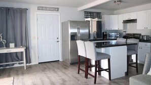 Una cocina moderna con un refrigerador de acero inoxidable, un microondas, un tostador y una cafetera. Hay una barra con dos taburetes y una pequeña mesa. La cocina tiene gabinetes blancos y una encimera gris.
