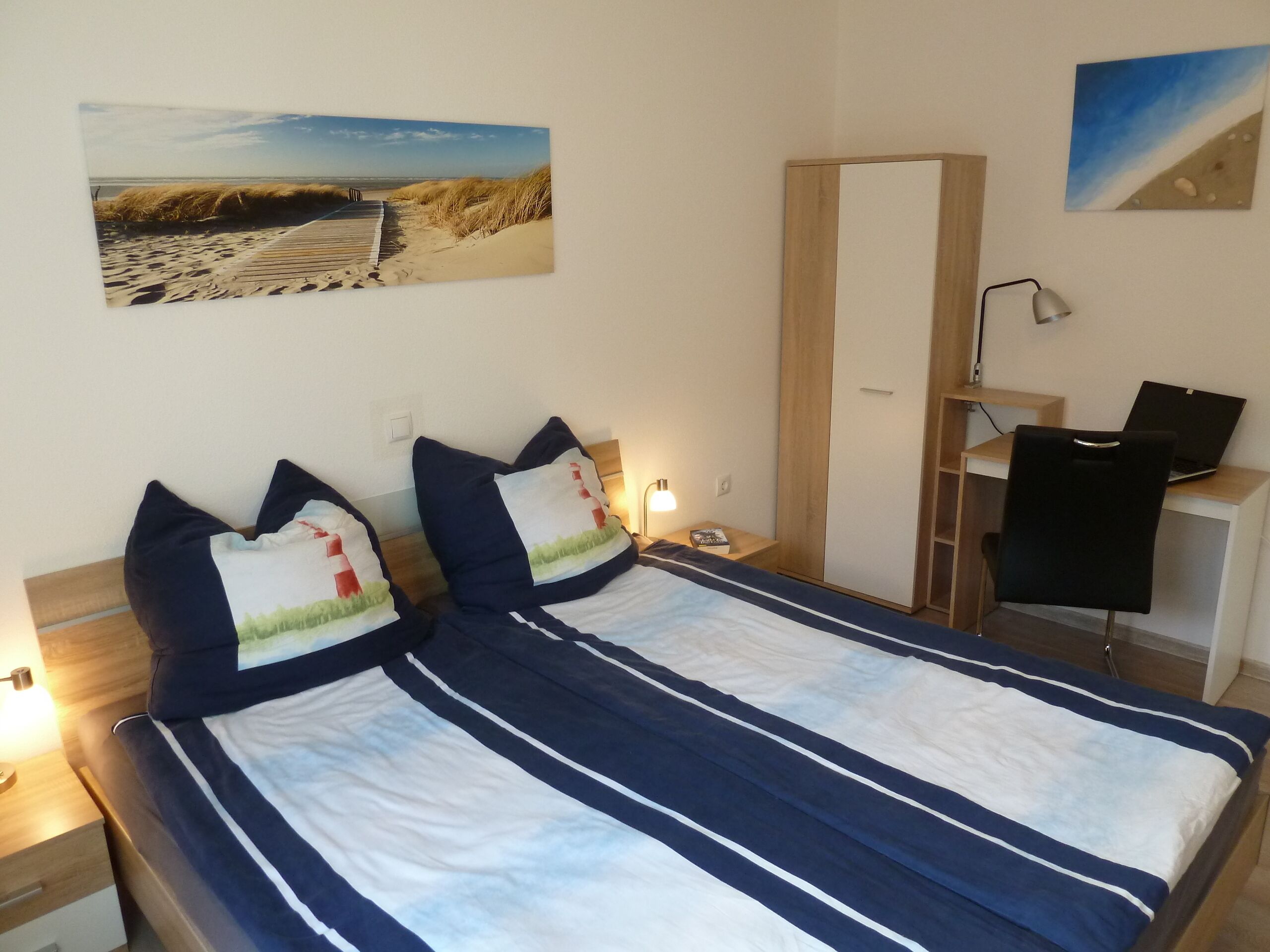 A double bed with striped bedding, a desk with a chair, a lamp, and two wall-mounted paintings.