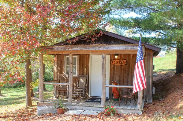A rustic cabin with a porch, a wooden sign reading "FOX DEN", and an American flag.