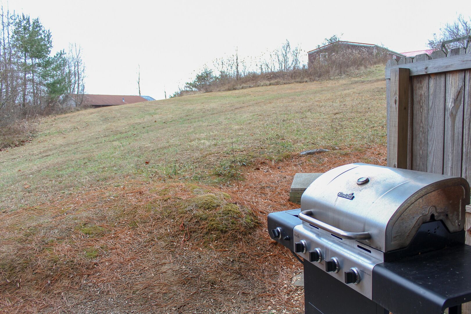 An outdoor grill with a control panel, set against a grassy hillside with a wooden structure in the background.