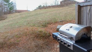 Una parrilla al aire libre con panel de control, frente a una ladera cubierta de césped y una estructura de madera al fondo.
