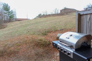 An outdoor grill with a control panel, set against a grassy hillside with a wooden structure in the background.