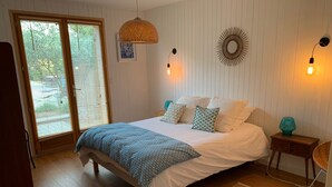 A bedroom with a bed, a wooden floor, a window with blinds, a wall-mounted light fixture, and a small table with a lamp. - Wooden house in the center of Cap Ferrets ans a quiet street 100 m from the market. (Lège-Cap-Ferret)